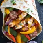 Sheet pan chicken and sausage and pepper wrap - the image shows a tortilla wrap filled with a variety of colorful vegetables. the wrap is made of tortilla and is filled with grilled chicken, red, yellow, green, and orange bell peppers, sliced red onions, and sliced red and yellow bell peppers. the chicken is cooked to a golden brown and appears to be seasoned with herbs and spices. there are also a few sprigs of cilantro scattered around the wrap, adding a pop of green to the dish. the background is a dark black slate surface.