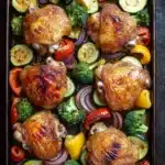 Sheet pan chicken and veggies - the image shows a rectangular baking tray filled with roasted chicken and vegetables. the chicken is golden brown and appears to be seasoned with herbs and spices. the vegetables include zucchini, broccoli, red onions, and yellow bell peppers. the tray is placed on a dark wooden table. the overall presentation of the dish is colorful and appetizing.
