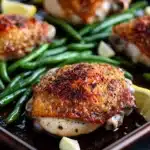 Sheet pan chicken green beans - the image shows a baking tray with four pieces of grilled chicken breasts on it. the chicken breasts are golden brown and appear to be seasoned with herbs and spices. the tray is lined with green beans and there are lemon wedges scattered around the tray. the background is blurred, but it appears to be a kitchen countertop. the overall color scheme of the image is warm and inviting.