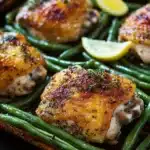Sheet pan chicken green beans - the image shows a tray of grilled chicken breasts with green beans. the chicken breasts are golden brown and appear to be seasoned with herbs and spices. the green beans are bright green and look fresh and vibrant. there are two lemon wedges on the side of the tray, adding a pop of color to the dish. the tray is lined with parchment paper, and the background is blurred, making the chicken and beans the focal point of the image.