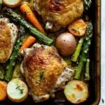Sheet pan chicken with roasted spring vegetables - the image shows a baking tray with roasted chicken and vegetables. the chicken is golden brown and appears to be seasoned with herbs and spices. the vegetables include asparagus, carrots, potatoes, and lemons. the tray is lined with parchment paper and there is a sprig of rosemary on the side. the background is a white marble countertop.