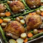 Sheet pan chicken with roasted spring vegetables - the image shows a baking tray filled with roasted chicken and vegetables. the chicken is golden brown and appears to be seasoned with herbs and spices. the vegetables include asparagus, carrots, potatoes, and peas. the tray is lined with parchment paper and is placed on a wooden table. the colors of the vegetables are vibrant and the dish looks appetizing.
