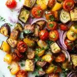 Sheet pan dinner eggplant - the image is a close-up of a colorful salad on a white marble surface. the salad is made up of grilled eggplant, cherry tomatoes, red onions, and yellow bell peppers. the eggplant is cut into small cubes and is arranged in a circular pattern. the cherry tomatoes are bright red and appear to be ripe and juicy. the red onions are sliced into thin strips and are scattered throughout the salad. there are also some chopped parsley scattered throughout, adding a pop of green color to the dish. the overall appearance is fresh and appetizing.