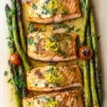 Sheet pan lemon brown butter salmon - the image shows a rectangular baking tray with four pieces of salmon and asparagus arranged in a diagonal line. the salmon is cooked to a golden brown color and appears to be seasoned with herbs and spices. the asparaglips are bright green and look fresh and vibrant. there are also small cherry tomatoes scattered around the tray, adding a pop of color to the dish. the tray is lined with a light-colored parchment paper, and there are a few sprigs of parsley scattered around it. the overall presentation of the dish is elegant and appetizing.