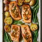 Sheet pan lemon brown butter salmon - the image shows a baking tray with four pieces of salmon on it. the salmon is cooked to a golden brown color and appears to be seasoned with herbs and spices. on top of the salmon, there are roasted potatoes and green beans. the potatoes are golden brown and have a crispy texture. the green beans are bright green and look fresh and vibrant. the tray is lined with parchment paper and is placed on a white marble countertop.