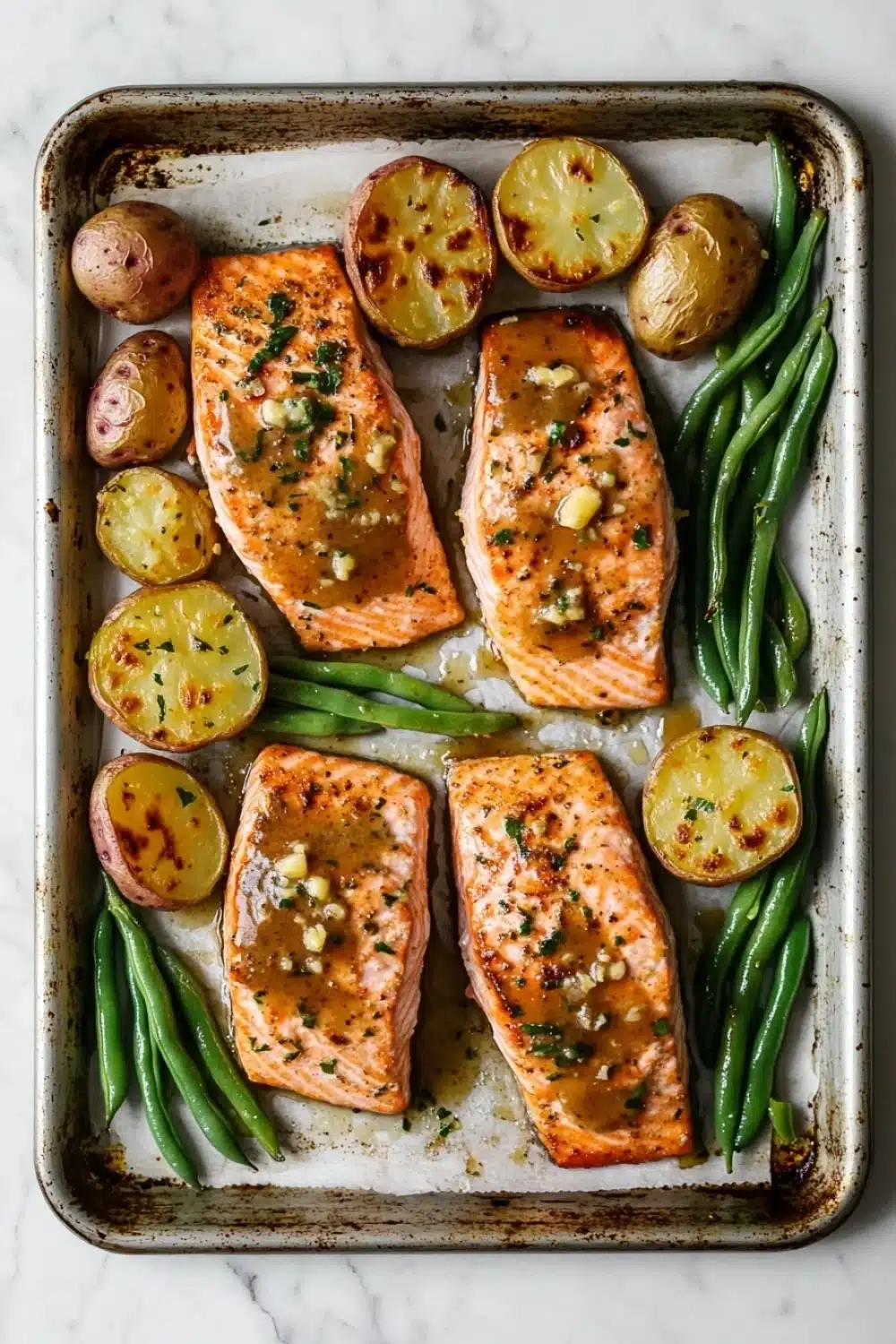 Sheet pan lemon brown butter salmon - the image shows a baking tray with four pieces of salmon on it. the salmon is cooked to a golden brown color and appears to be seasoned with herbs and spices. on top of the salmon, there are roasted potatoes and green beans. the potatoes are golden brown and have a crispy texture. the green beans are bright green and look fresh and vibrant. the tray is lined with parchment paper and is placed on a white marble countertop.