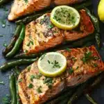 Sheet pan salmon green beans - the image shows two pieces of salmon fillets on a black slate plate. the salmon is cooked to a golden brown color and is garnished with fresh herbs and lemon wedges. the plate is surrounded by a bed of green asparagus spears. the background is blurred, but it appears to be a kitchen countertop. the overall presentation of the dish is elegant and appetizing.