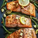 Sheet pan salmon green beans - the image shows two pieces of salmon fillets on a black baking tray. the salmon is cooked to a golden brown color and appears to be seasoned with herbs and spices. on top of the salmon, there are two slices of lemon and a few pieces of green beans scattered around. the green beans are bright green and look fresh and vibrant. the tray is lined with parchment paper, giving the image a rustic and appetizing feel.