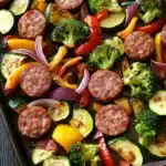 Sheet pan smoked sausage and veggies - the image shows a tray of cooked vegetables on a wooden table. the vegetables appear to be zucchini, broccoli, red and yellow bell peppers, and sausage. the sausage is cut into small cubes and is arranged in a circular pattern on the tray. the colors of the vegetables are vibrant and the dish looks appetizing. the tray is lined with parchment paper and the background is dark, making the vegetables stand out.