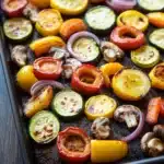 Sheet pan vegetables air fryer - the image shows a baking tray filled with roasted vegetables. the vegetables are arranged in a circular pattern on the tray, with some overlapping each other. the colors of the vegetables include red, yellow, green, and orange bell peppers, as well as sliced red onions and mushrooms. the tray is lined with parchment paper, and the vegetables appear to be seasoned with herbs and spices. the background is a wooden table.