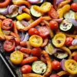 Sheet pan vegetables air fryer - the image shows a tray of roasted vegetables on a wooden table. the vegetables are arranged in a circular pattern on the tray, with some overlapping each other. the colors of the vegetables include red, yellow, and orange bell peppers, zucchini, and cherry tomatoes. there are also sliced red onions scattered throughout the vegetables. the tray appears to be made of metal and has a shiny surface. the background is blurred, but it seems to be a kitchen countertop.