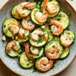 Shrimp and zucchini skillet - the image is a close-up of a plate of shrimp and zucchini salad. the plate is round and has a light blue speckled pattern. the shrimp are arranged in a circular pattern on top of the zucchinis, which are cut into small cubes and are garnished with chopped parsley. the dish is served on a white tablecloth with a few sprigs of parsley scattered around the plate. the overall color scheme of the dish is warm and inviting.