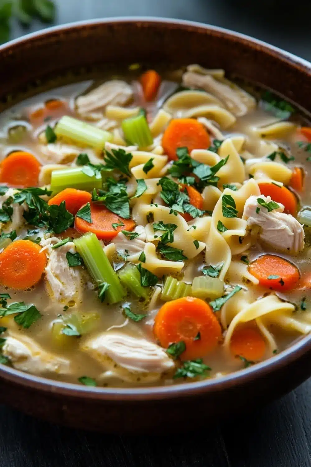 Skillet chicken noodle soup - the image is a close-up of a bowl of chicken noodle soup. the soup is made with noodles, carrots, celery, and chunks of chicken. the broth is a light beige color and appears to be thick and creamy. the bowl is made of dark brown ceramic and is sitting on a dark wooden table. there are a few sprigs of parsley scattered throughout the soup, adding a pop of green color to the dish. the background is blurred, making the soup the focal point of the image.