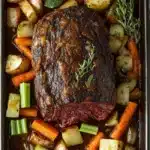 Slow cooker beef chuck roast - the image shows a rectangular baking dish filled with a roast beef dish. the roast beef is cooked medium-rare and has a rich, dark brown color. it is garnished with fresh herbs, including rosemary sprigs and thyme. the dish is served on a bed of diced potatoes, carrots, celery, and other vegetables. the potatoes are golden brown and appear to be seasoned with herbs and spices. the carrots are bright orange and are cut into small cubes. the celery is a bright green color and is scattered throughout the dish. there is a blue and white striped napkin on the side of the dish, adding a pop of color to the image.