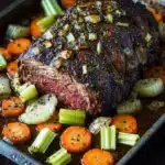 Slow cooker beef chuck roast - the image shows a large piece of cooked meat on a baking tray. the meat appears to be a medium-rare ribeye steak, with a dark brown crust on top. it is garnished with chopped onions and celery, and there are also small pieces of carrots and onions scattered around the dish. the tray is sitting on a wooden table, and the background is blurred, making the meat the focal point of the image.