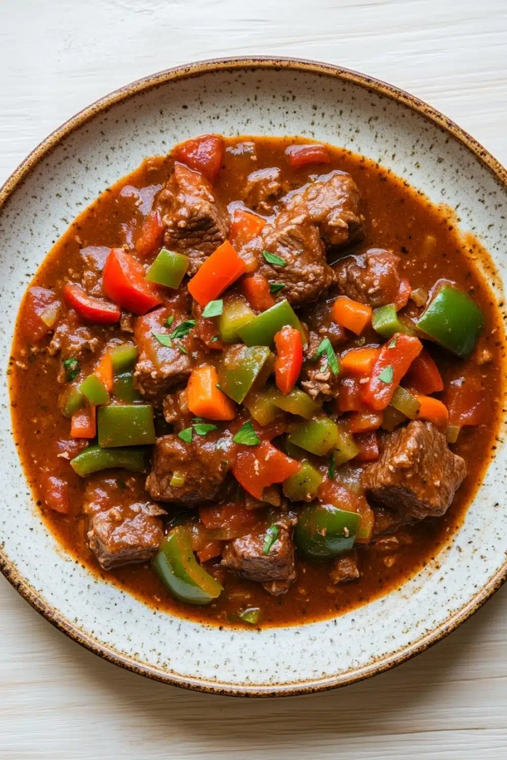 Slow cooker beef goulash - the image is a close-up of a dish in a bowl. the dish appears to be a stew or stew with chunks of beef and vegetables mixed in. the beef is cooked in a tomato-based sauce and is garnished with chopped red and green bell peppers. the vegetables are cut into small pieces and are scattered throughout the dish. the bowl is white with a speckled pattern around the edges. the background is a light-colored wooden table.