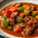 Slow cooker beef goulash - the image is a close-up of a bowl of beef stew. the stew is a rich orange color and appears to be made with chunks of beef, carrots, and bell peppers. the beef is cooked in a tomato-based sauce and is garnished with chopped onions. the vegetables are cut into small pieces and are mixed in with the beef. the bowl is white with a blue rim and is sitting on a dark blue tablecloth.