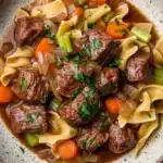 Slow cooker beef noodle - the image is a close-up of a bowl of beef noodle soup. the soup is made with chunks of beef, carrots, celery, and onions, and is garnished with chopped parsley. the bowl is made of ceramic with a speckled pattern and is sitting on a white marble countertop. the broth is a rich brown color and appears to be thick and creamy. the noodles are long and thin, and the vegetables are bright orange and green. the dish looks appetizing and ready to eat.