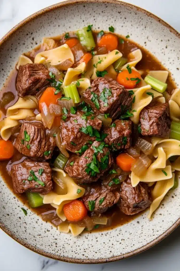 Slow cooker beef noodle - the image is a close-up of a bowl of beef noodle soup. the soup is made with chunks of beef, carrots, celery, and onions, and is garnished with chopped parsley. the bowl is made of ceramic with a speckled pattern and is sitting on a white marble countertop. the broth is a rich brown color and appears to be thick and creamy. the noodles are long and thin, and the vegetables are bright orange and green. the dish looks appetizing and ready to eat.
