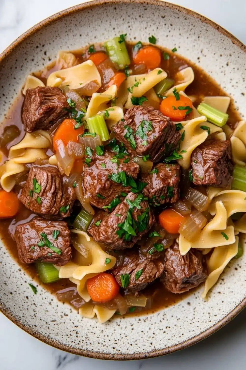 Slow cooker beef noodle - the image is a close-up of a bowl of beef noodle soup. the soup is made with chunks of beef, carrots, celery, and onions, and is garnished with chopped parsley. the bowl is made of ceramic with a speckled pattern and is sitting on a white marble countertop. the broth is a rich brown color and appears to be thick and creamy. the noodles are long and thin, and the vegetables are bright orange and green. the dish looks appetizing and ready to eat.