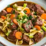 Slow cooker beef noodle - the image is a close-up of a bowl of beef stew. the stew is made with chunks of beef, carrots, celery, and noodles. the beef is cooked in a rich, dark brown broth and is garnished with chopped parsley. the noodles are long and thin, and the carrots are bright orange. the dish is served in a light blue bowl with a white rim. the bowl is sitting on a white marble countertop.