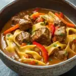 Slow cooker beef noodle curry - the image is a close-up of a bowl of beef noodle soup. the bowl is made of ceramic and has a speckled pattern on the sides. the soup is a rich orange color and appears to be thick and creamy. it is filled with chunks of beef, red bell peppers, and sliced carrots. the noodles are thin and yellow, and the sauce is thick and thick. there are also small pieces of green herbs scattered throughout the soup, adding a pop of color to the dish. the background is a dark grey countertop.