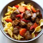 Slow cooker beef noodle curry - the image is a close-up of a bowl of beef stew. the bowl is made of dark brown ceramic and is sitting on a white wooden table. the stew is made up of chunks of beef, carrots, and noodles. the beef is cooked in a rich, dark brown sauce and appears to be seasoned with herbs and spices. the noodles are long and thin, and they are arranged in a pile on top of the beef. the colors of the stew are vibrant and the dish looks appetizing.