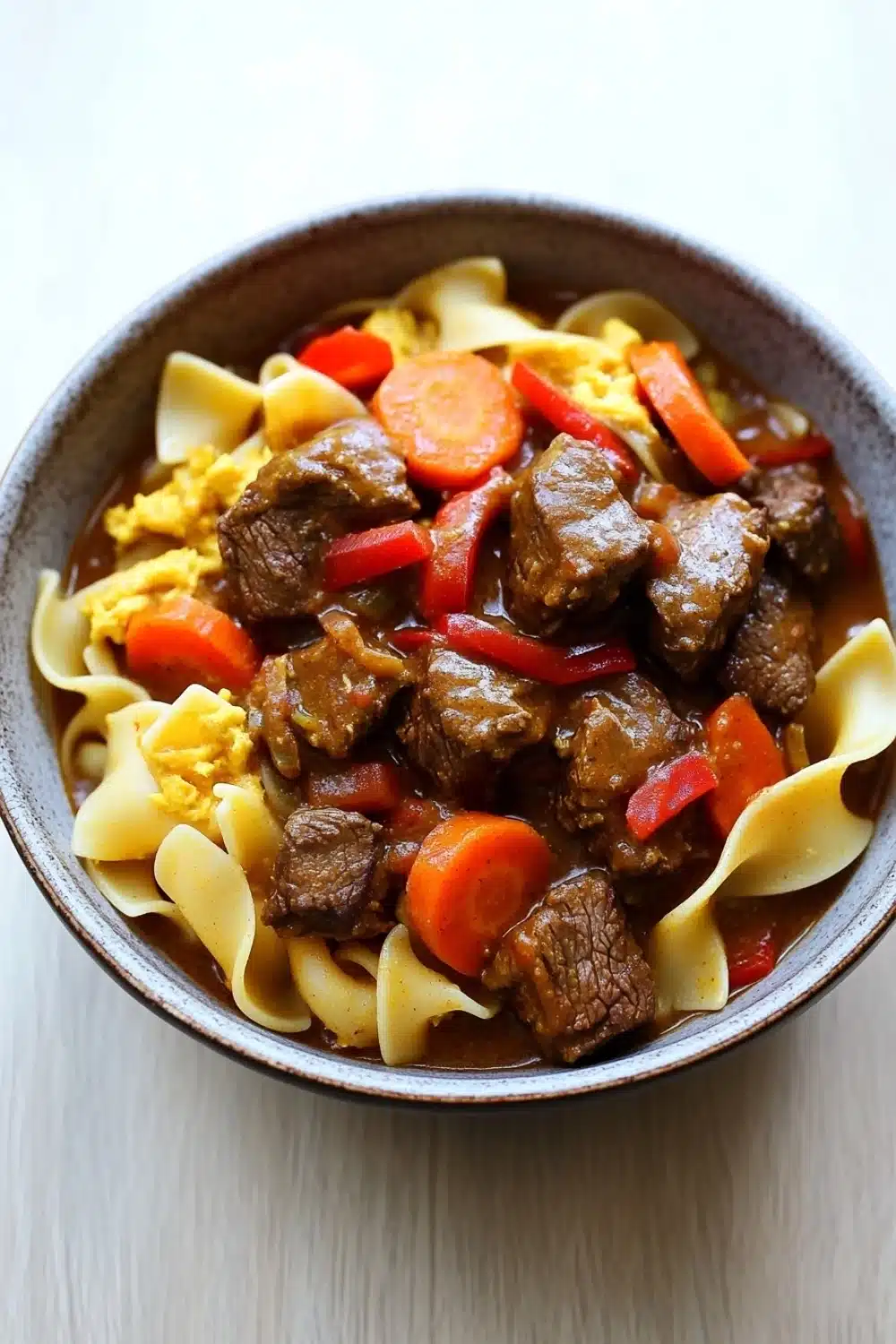 Slow cooker beef noodle curry - the image is a close-up of a bowl of beef stew. the bowl is made of dark brown ceramic and is sitting on a white wooden table. the stew is made up of chunks of beef, carrots, and noodles. the beef is cooked in a rich, dark brown sauce and appears to be seasoned with herbs and spices. the noodles are long and thin, and they are arranged in a pile on top of the beef. the colors of the stew are vibrant and the dish looks appetizing.