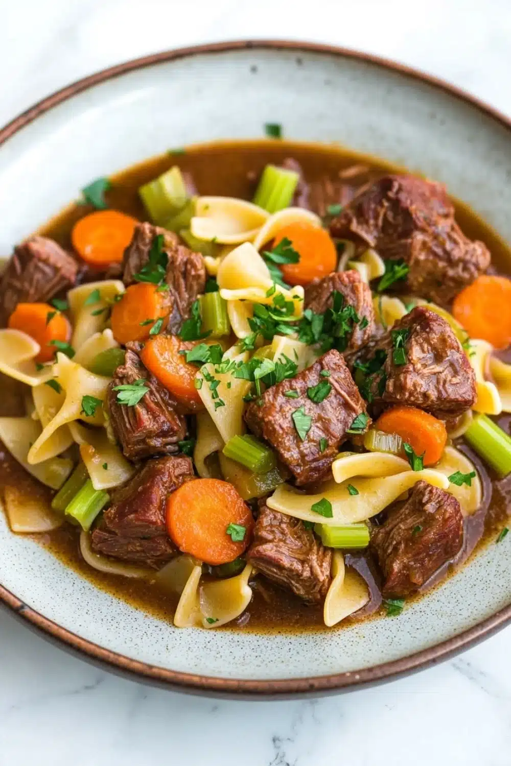 Slow cooker beef noodle - the image is a close-up of a bowl of beef stew. the stew is made with chunks of beef, carrots, celery, and noodles. the beef is cooked in a rich, dark brown broth and is garnished with chopped parsley. the noodles are long and thin, and the carrots are bright orange. the dish is served in a light blue bowl with a white rim. the bowl is sitting on a white marble countertop.