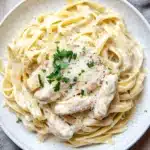 Slow cooker chicken alfredo - the image shows a plate of fettuccine alfredo pasta with a creamy white sauce and chunks of chicken. the pasta is cooked al dente and is topped with a sprinkle of parmesan cheese and fresh parsley. the plate is white and is sitting on a wooden table with a gray cloth napkin on the side. there are also a few sprigs of parsley scattered around the plate. the overall presentation of the dish is elegant and appetizing.