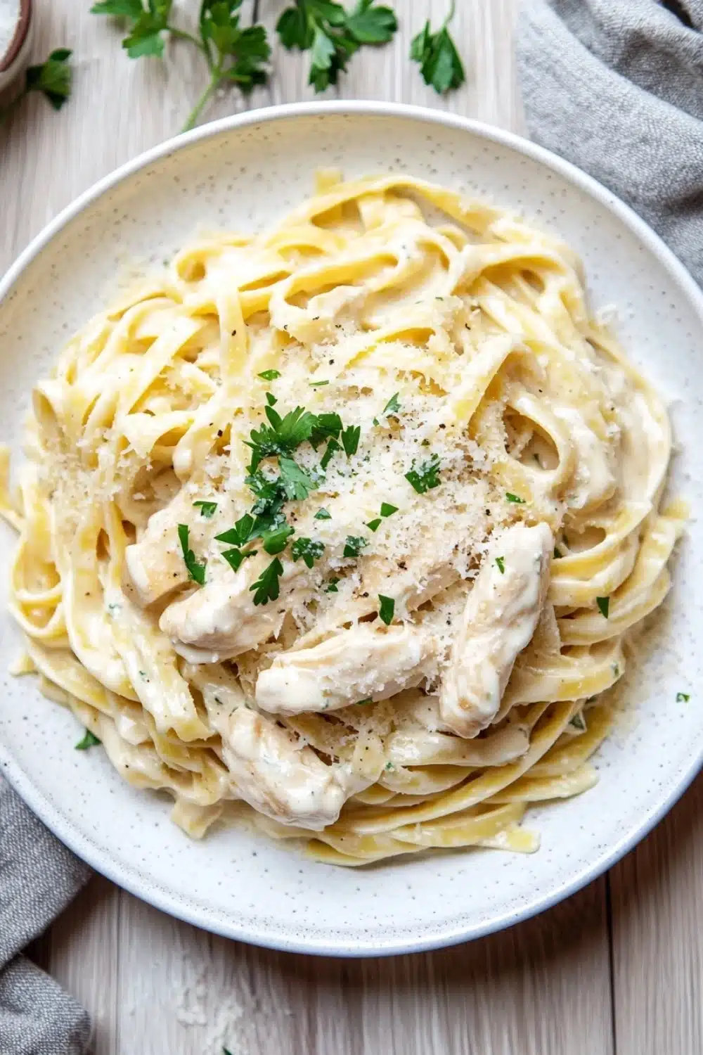 Slow cooker chicken alfredo - the image shows a plate of fettuccine alfredo pasta with a creamy white sauce and chunks of chicken. the pasta is cooked al dente and is topped with a sprinkle of parmesan cheese and fresh parsley. the plate is white and is sitting on a wooden table with a gray cloth napkin on the side. there are also a few sprigs of parsley scattered around the plate. the overall presentation of the dish is elegant and appetizing.