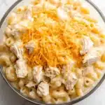 Slow cooker chicken mac and cheese - the image is a close-up of a bowl of macaroni and cheese. the bowl is made of glass and is sitting on a white marble countertop. the cheese is a bright yellow color and appears to be melted and bubbly. on top of the cheese, there are chunks of chicken that are scattered throughout the dish. the chicken is a light brown color and looks crispy on the outside. the dish looks creamy and ready to eat.