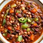 Slow cooker chili vegan - the image is a close-up of a bowl of chili. the chili is a rich orange color and appears to be made with beans, carrots, and other vegetables. the beans are black and white, and the carrots are bright orange. there are also some green bell peppers scattered throughout the chili, adding a pop of color to the dish. the bowl is sitting on a white marble countertop.