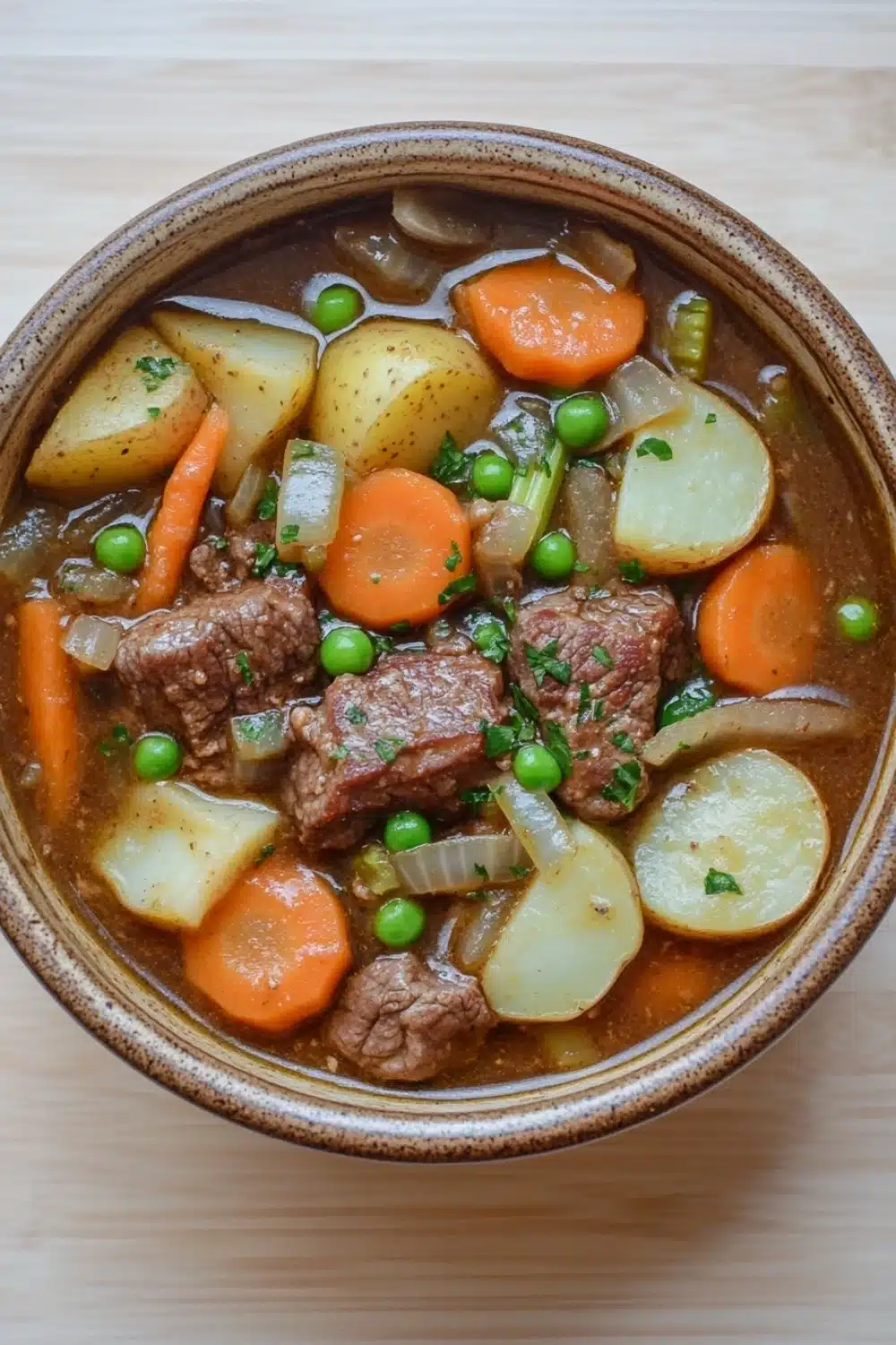Slow cooker old fashion beef stew - the image is a close-up of a bowl of beef stew. the stew is made with chunks of beef, carrots, potatoes, and peas. the beef is cooked medium-rare and appears to be seasoned with herbs and spices. the potatoes are cut into small cubes and are arranged in a circular pattern around the beef. the carrots are bright orange and the peas are green. the broth is a rich brown color and looks thick and creamy. the bowl is made of ceramic and is sitting on a wooden table.