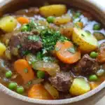 Slow cooker old fashion beef stew - the image is a close-up of a bowl of beef stew. the stew is made with chunks of beef, carrots, potatoes, and peas. the beef is cooked in a rich, dark brown broth and is garnished with chopped parsley. the potatoes are yellow and appear to be seasoned with herbs and spices. the carrots are bright orange and the peas are green. the bowl is made of ceramic and is sitting on a wooden table. the background is blurred, but it appears to be a kitchen countertop.