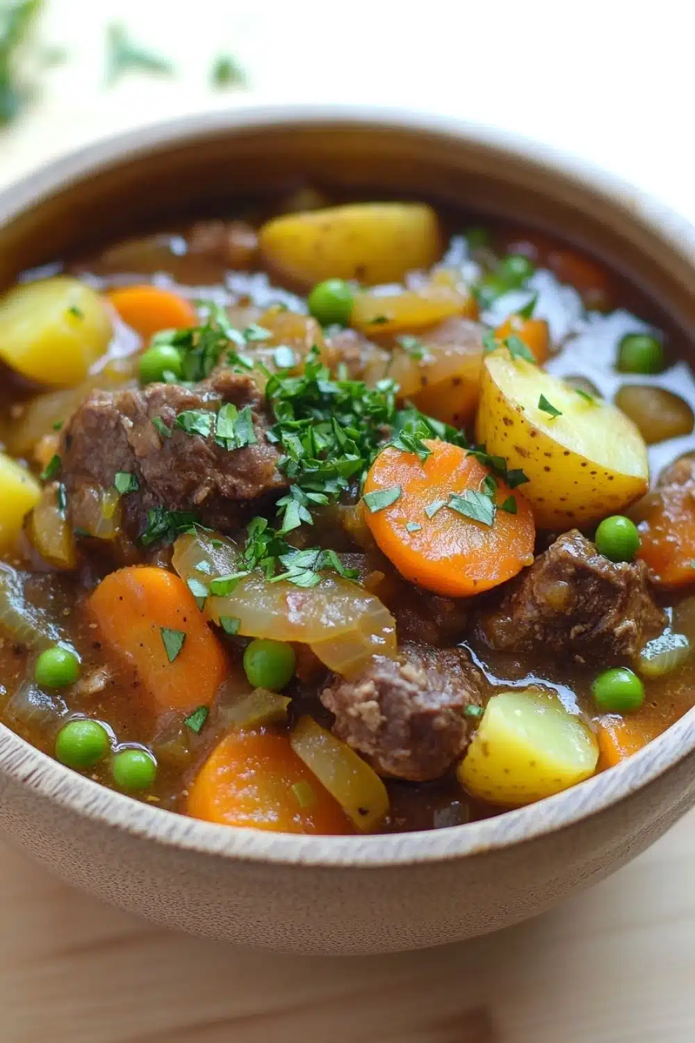 Slow cooker old fashion beef stew - the image is a close-up of a bowl of beef stew. the stew is made with chunks of beef, carrots, potatoes, and peas. the beef is cooked in a rich, dark brown broth and is garnished with chopped parsley. the potatoes are yellow and appear to be seasoned with herbs and spices. the carrots are bright orange and the peas are green. the bowl is made of ceramic and is sitting on a wooden table. the background is blurred, but it appears to be a kitchen countertop.