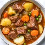 Slow cooker poor mans beef stew - the image is a close-up of a bowl of beef stew. the stew is made with chunks of beef, potatoes, carrots, peas, and onions. the beef is cooked medium-rare and appears to be seasoned with herbs and spices. the potatoes are yellow and the carrots are bright orange. the peas are green and appear to be peas. the broth is a rich orange color and is thick and thick. the bowl is white with a speckled rim. the background is a white marble countertop.