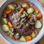 Slow cooker pot roast with cream of mushroom soup - the image is a close-up of a bowl of beef stew. the stew is made with chunks of beef, mushrooms, carrots, potatoes, and onions. the beef is cooked medium-rare and appears to be seasoned with herbs and spices. the potatoes are yellow and the carrots are orange. the onions are red and are scattered throughout the stew, adding a pop of color to the dish. the bowl is made of ceramic and is sitting on a wooden table.