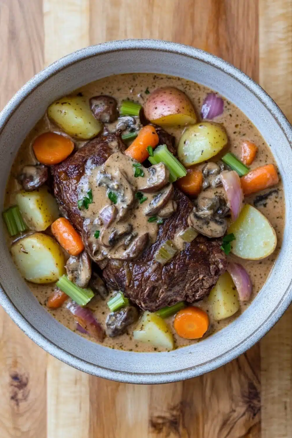 Slow cooker pot roast with cream of mushroom soup - the image is a close-up of a bowl of beef stew. the stew is made with chunks of beef, mushrooms, carrots, potatoes, and onions. the beef is cooked medium-rare and appears to be seasoned with herbs and spices. the potatoes are yellow and the carrots are orange. the onions are red and are scattered throughout the stew, adding a pop of color to the dish. the bowl is made of ceramic and is sitting on a wooden table.
