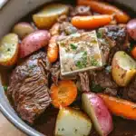 Slow cooker pot roast with ranch packet - the image is a close-up of a pot of beef stew. the pot is made of stainless steel and is sitting on a wooden table. the stew is made up of chunks of beef, carrots, potatoes, and onions. the beef is cooked medium-rare and appears to be seasoned with herbs and spices. there is a small piece of bread on top of the stew, which is covered in a dark brown sauce. the vegetables are arranged in a circular pattern around the beef. the overall appearance of the dish is hearty and flavorful.