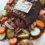 Slow cooker pot roast with ranch packet - the image shows a large piece of roast beef on a wooden cutting board. the roast beef is cooked medium-rare and has a rich, dark brown color. it is garnished with sliced carrots, onions, and potatoes. the potatoes are cut into small cubes and are arranged around the roast beef. there is a label on top of the roast that reads "roast beef". the dish appears to be freshly cooked and ready to eat.
