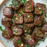 Steak bites with garlic butter crockpot - the image shows a plate of cooked steak cubes. the steak cubes are arranged in a circular pattern on a light blue plate with a white rim. they are covered in a dark brown sauce and garnished with chopped parsley. the dish appears to be seasoned with herbs and spices, giving it a savory flavor. the plate is sitting on a wooden table with a light beige background.