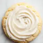 Sugar cookie and frosting - the image shows a round cookie with a swirl of white frosting on top. the cookie appears to be freshly baked and is placed on a white marble surface. the frosting is in the shape of a spiral and is spread evenly across the cookie. the background is blurred, but it seems to be a kitchen countertop.