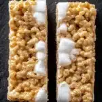 Sugar cookie rice crispy - the image shows two rectangular bars of rice krispy treats on a black slate background. the bars are golden brown in color and have a crumbly texture. they are topped with a generous amount of white sugar, which is drizzled over the top of each bar. the sugar appears to be sugar-free and has a smooth texture. the background is a solid black color, making the bars stand out.