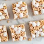 Sugar cookie rice crispy - the image shows a group of square-shaped rice krispy treats arranged on a white marble surface. the treats are made with oats, nuts, and marshmallows, and are arranged in a scattered manner. some of the treats have a golden-brown color, while others have a light brown color. there are also a few scattered nuts around the treats, adding texture and crunchy texture. the background is blurred, but it appears to be a kitchen countertop.