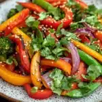 Thai vegetable air fryer - the image is a close-up of a colorful salad on a white plate with a speckled pattern. the salad is made up of sliced red and yellow bell peppers, red onions, and green bell peppers. the peppers are arranged in a circular pattern, with some overlapping each other. there are also some sesame seeds sprinkled on top. the plate is sitting on a wooden table with a white napkin in the background. the overall color scheme of the salad is vibrant and appetizing.