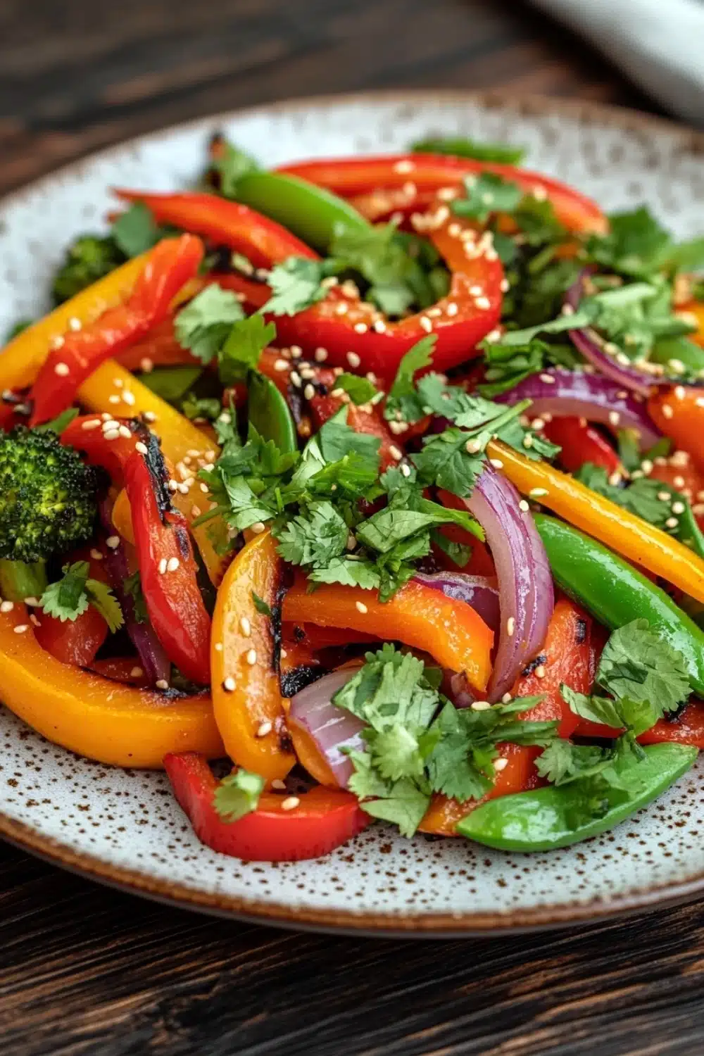 Thai vegetable air fryer - the image is a close-up of a colorful salad on a white plate with a speckled pattern. the salad is made up of sliced red and yellow bell peppers, red onions, and green bell peppers. the peppers are arranged in a circular pattern, with some overlapping each other. there are also some sesame seeds sprinkled on top. the plate is sitting on a wooden table with a white napkin in the background. the overall color scheme of the salad is vibrant and appetizing.