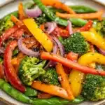 Thai vegetable air fryer - the image is a close-up of a plate of stir-fry dish. the dish is made up of various vegetables, including broccoli, red bell peppers, and green beans. the vegetables are arranged in a colorful and appetizing manner, with some overlapping each other. the colors of the vegetables are vibrant and the dish appears to be fresh and healthy. the plate is made of a light-colored ceramic or porcelain material and is sitting on a white marble countertop.