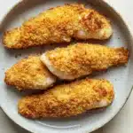 Tilapia in air fryer - the image shows a plate of four pieces of breaded fish fillets. the fillets are golden brown and appear to be crispy on the outside. they are arranged in a circular pattern on the plate, with the largest piece in the center and two smaller pieces on either side. the plate is light blue in color and has a textured surface. the background is a white wooden table.