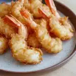 Trader joes shrimp tempura air fryer - the image shows a plate of fried shrimp on a wooden table. the shrimp are golden brown and appear to be crispy on the outside. they are arranged in a circular pattern on the plate, with some overlapping each other. the plate is brown with a speckled pattern around the edges. the background is blurred, but it appears to be a kitchen countertop.